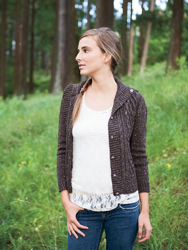 Freeport Cardigan - a knitwear pattern by Tian Connaughton for Knit Picks. A woman is shown standing in a wooded area wearing a brown knitted cardigan.