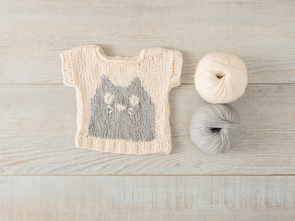 Knitted baby sweater with a cat face pattern in cream and gray using Knit Picks Snuggle Puff yarn and Claire Slade's Mini Meow sweater pattern next to 2 balls of yarn