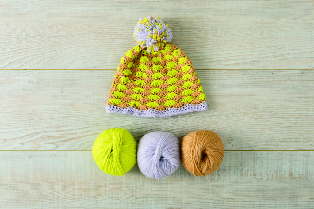 Yellow, tan and lavender crocheted hat with a pom-pom and shell stitches above three balls of Snuggle Puff yarn