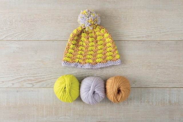 Yellow, tan and lavender crocheted hat with a pom-pom and shell stitches above three balls of Snuggle Puff yarn