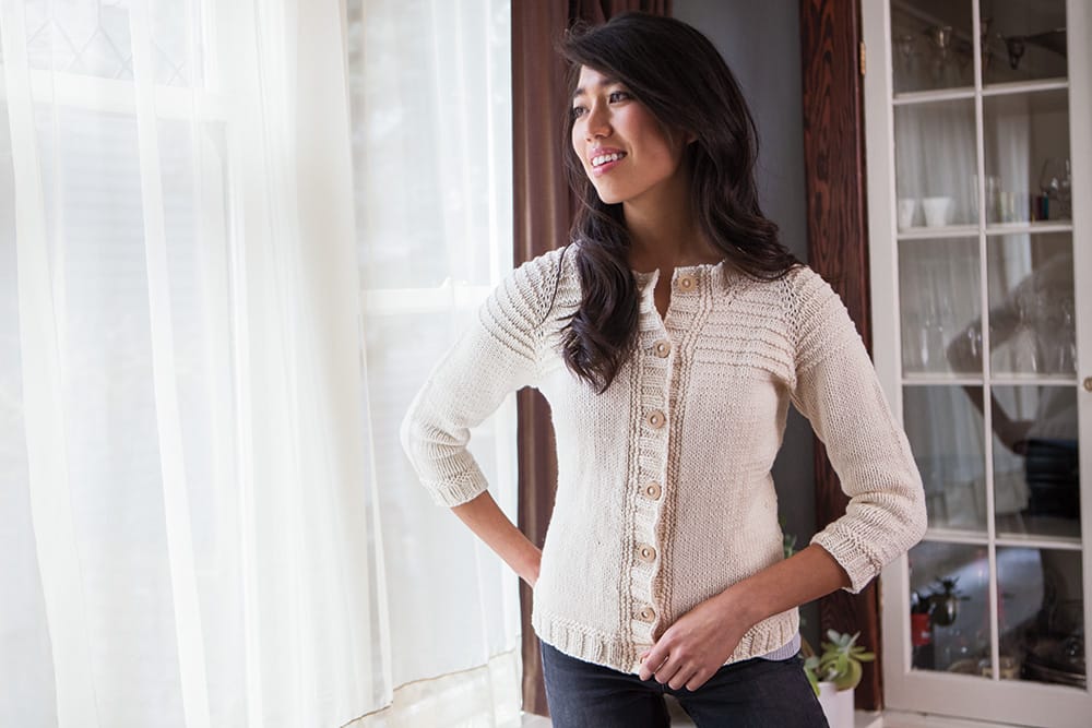 A woman wearing a natural-colored knit cardigan stands by a window