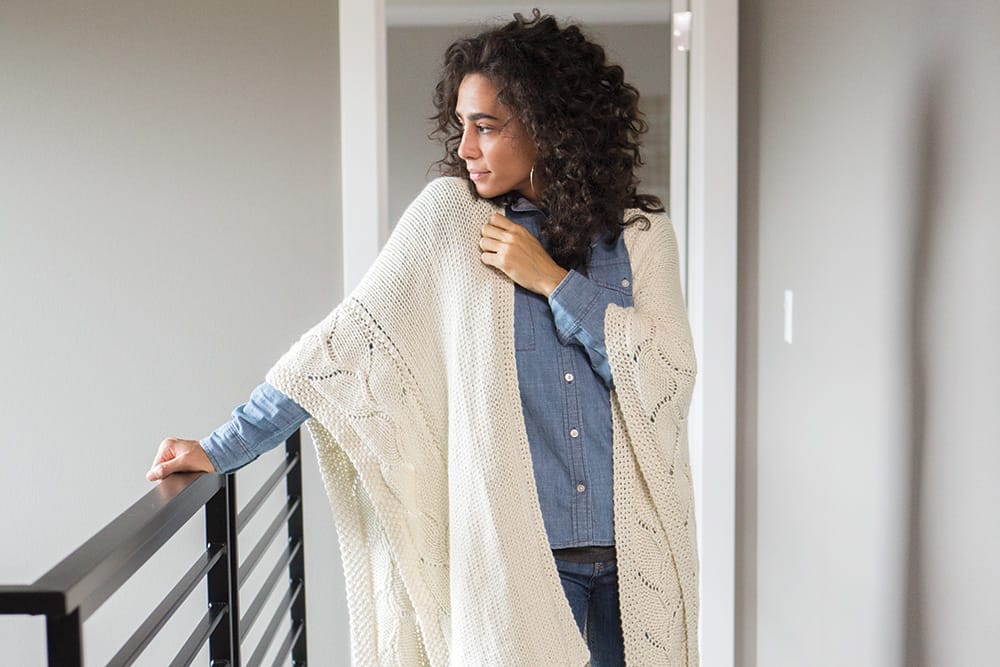 A woman wearing an oversized natural-colored poncho stands next to a railing