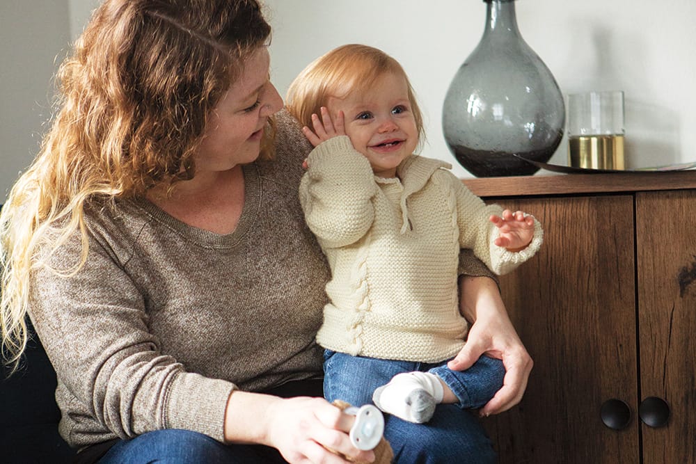 A mother holds a baby, who is wearing a cream-colored knitted pullover sweater