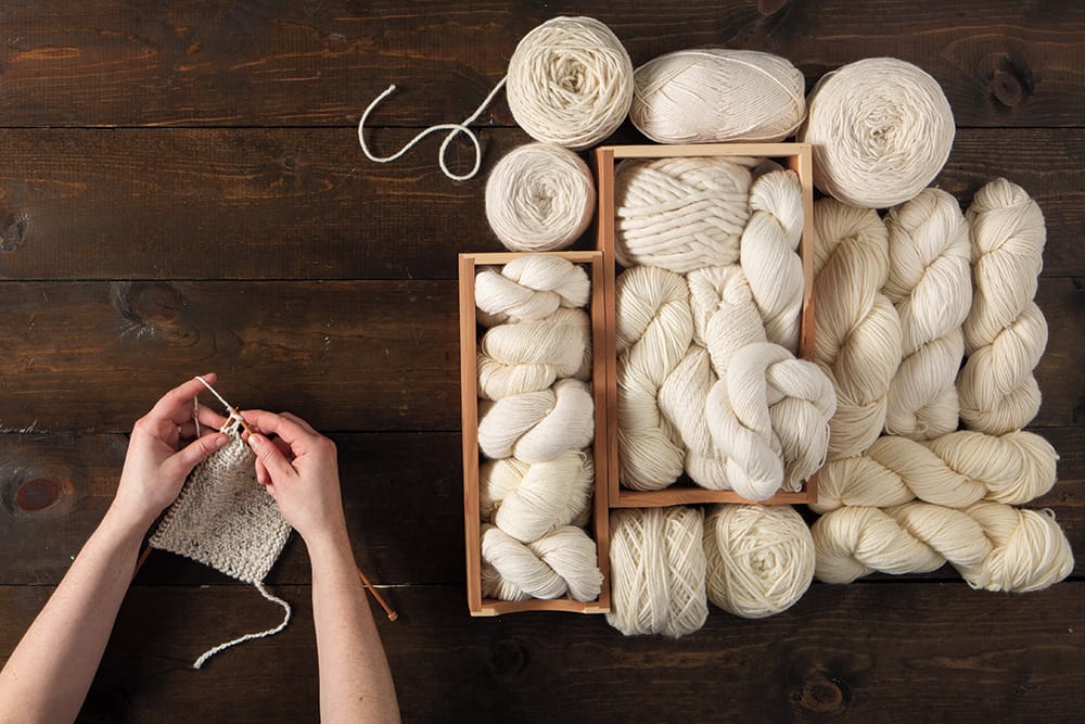 Hands knitting cream-colored yarn next to a pile of assorted undyed yarns in boxes.