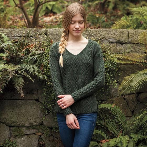 The Rhiannon Sweater, designed by Jenny Williams. A model stands against a stone wall wearing a green cabled sweater.