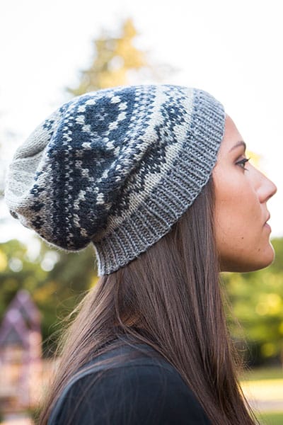 A model standing in profile wears the Telluride Slouch hat, a knitted hat designed by Jenny Williams using fair isle strand work patterning in gray, white, and blue.
