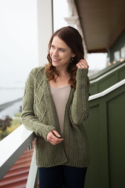 The McKenna Cardigan, designed by Jenny Williams. A model stands on a porch wearing a green knitted cardigan with cables.