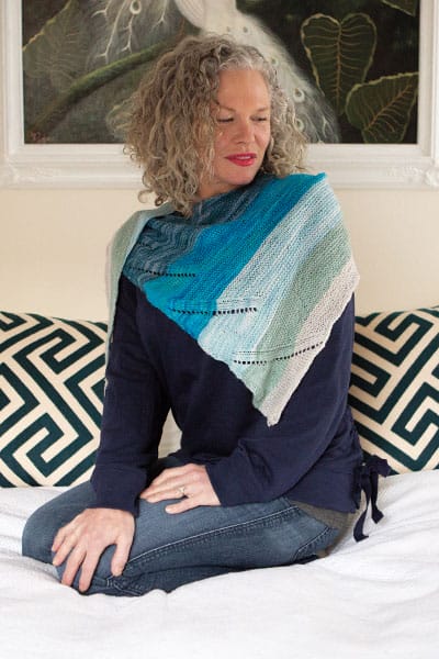 The Breakers Shawlette by Jenny Williams. A model sits on a bed wearing a knitted shawl with a variety of blue shades fading across it.