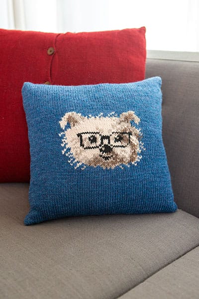 Clever Canine Cushion is a knitted blue pillow featuring a cute picture of a terrier dog face wearing black glasses.