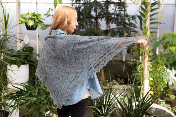 The Jardins Des Fleurs shawl. A model stands with her back to the camera, holding her arm out to display a lacy gray shawl.