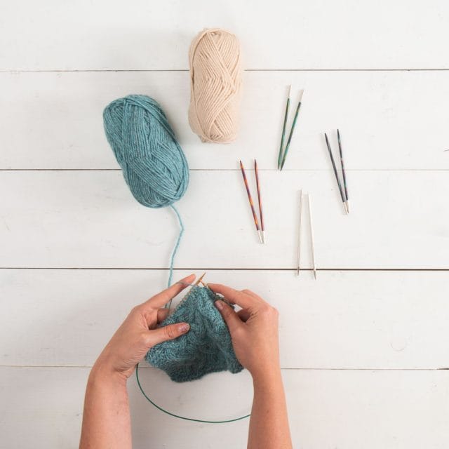 A top-down view of hands knitting from a ball of blue yarn. Another ball of white yarn sits on the table, next to four pairs of interchangeable knitting needles.