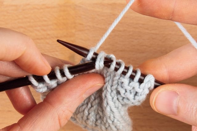 An image of two hands knitting with knitting needles and gray yarn