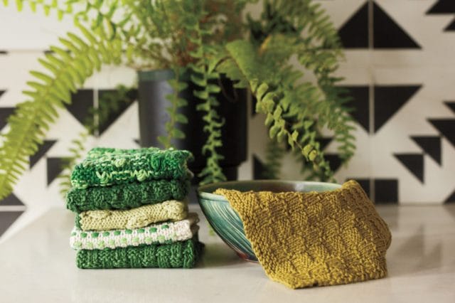 A stack of green dishcloths on a table top. One knitted dishcloth is draped in a green bowl.