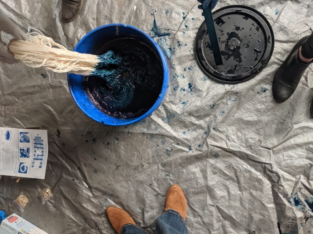 A top-down view of our indigo dyeing area. A silver tarp on the floor, a blue bucket full of indigo liquid, a skein of bare yarn being dipped into the bucket, and a stir stick.