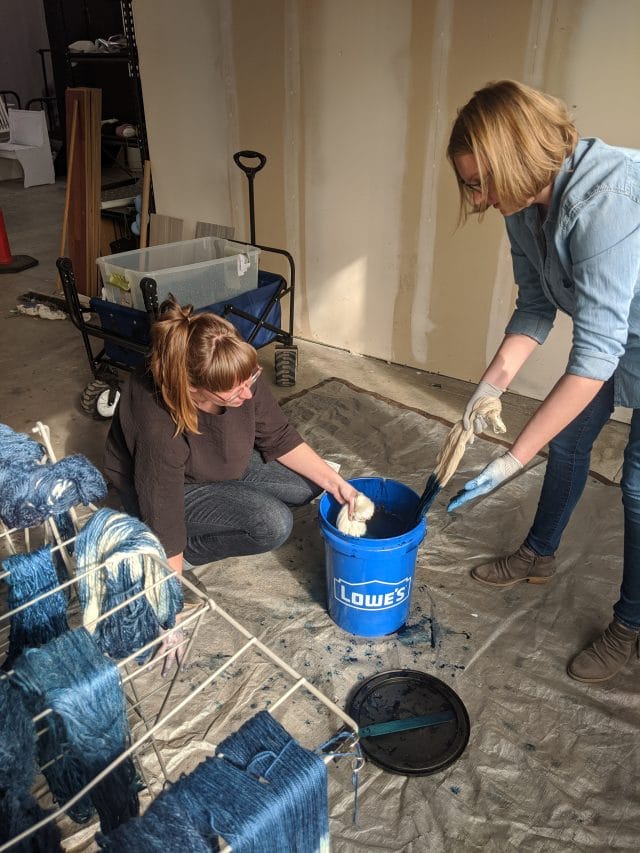 A photo of our indigo dyeing area: floor covered with a tarp, a bin of clear water in a rolling cart in the background. A woman crouches on the ground next to a 5 gallon bucket filled with indigo dye, and dips a skein of yarn inside while another woman stands nearby with a skein of yarn she is dyeing. In the foreground, a rack of drying yarn.