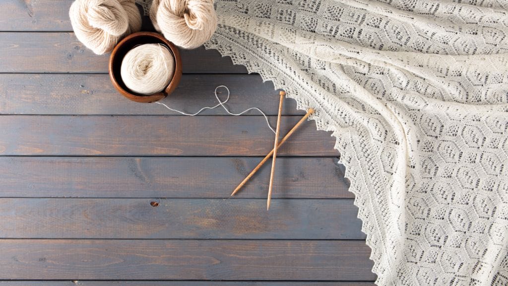 A wooden plank background with natural-colored yarn hanks and yarn in a yarn bowl at the top. A natural colored shawl drapes artfully across the background, with natural wood knitting needles crossed in the middle.