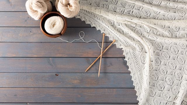 A wooden plank background with natural-colored yarn hanks and yarn in a yarn bowl at the top. A natural colored shawl drapes artfully across the background, with natural wood knitting needles crossed in the middle.