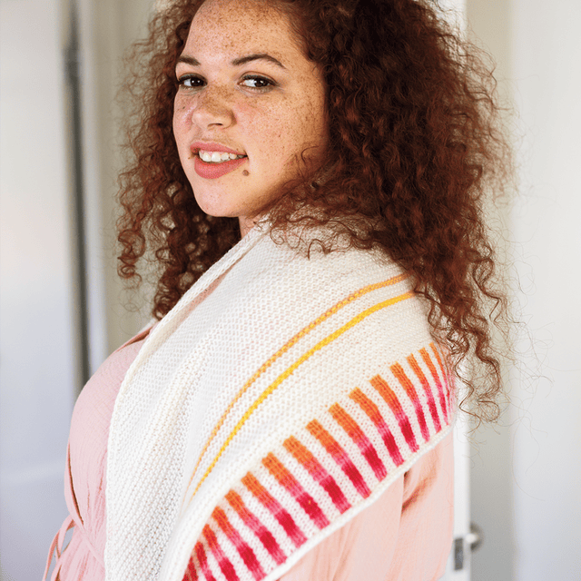 A model wears a cream knitted shaw with yellow-orange-pink-red striped accents.
