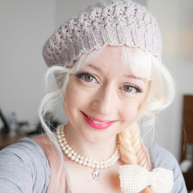 A headshot of Violet LeBeaux. She's wearing a knitted beret, a pearl necklace with a crown charm, and a pearl hair bow in her blonde braid.