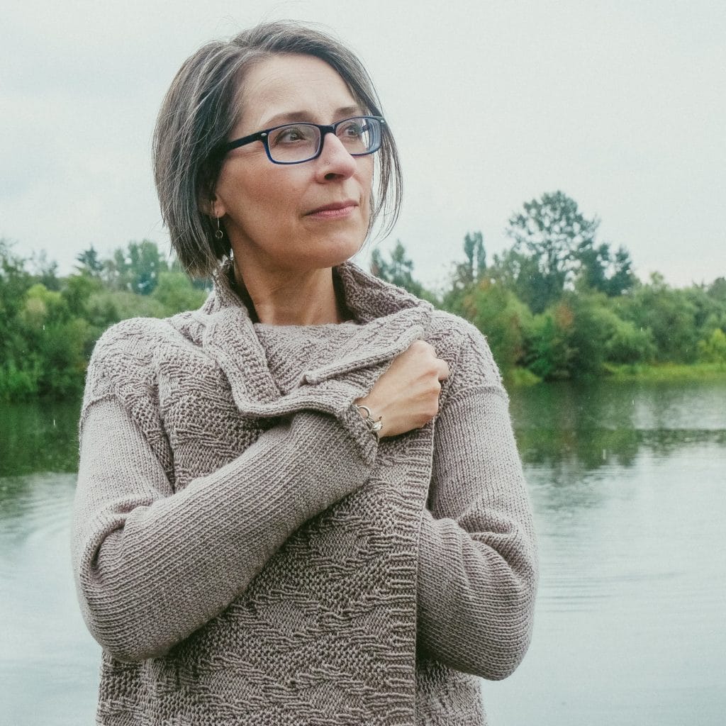 A headshot of Holli Yeoh, looking to the right and snuggling up in a pretty taupe knitted cardigan with a diamond pattern.