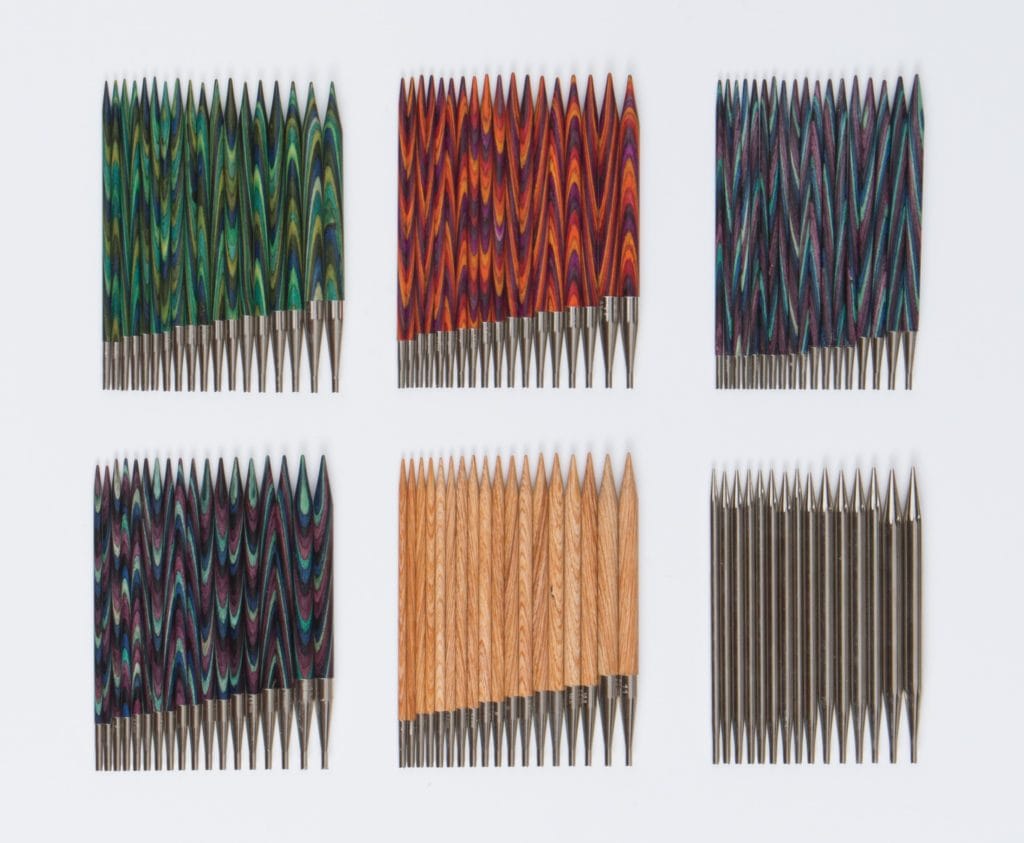 On a white background, six sets of interchangeable knitting needles: Green Caspian needles, Red/Purple Radiant needles, Two sets of Purple/Blue Majestic needles, blonde wood Sunstruck needles, and gray metal Nickel needles.