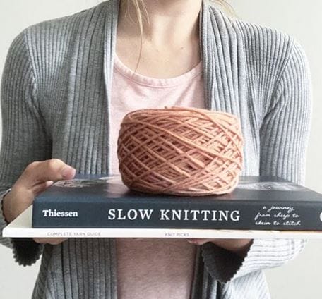 A person holds two knitting books with a cake of pinky-tan yarn on top.