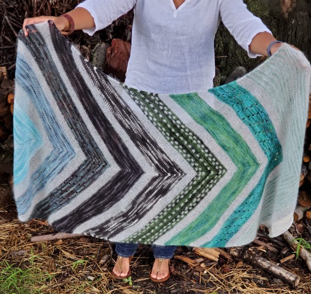 A woman holds up a knitted shawl in multicolored green, black, and gray chevrons. Kaliya Wrap by Tamy Gore: A new indie knitting pattern available at knitpicks.com.