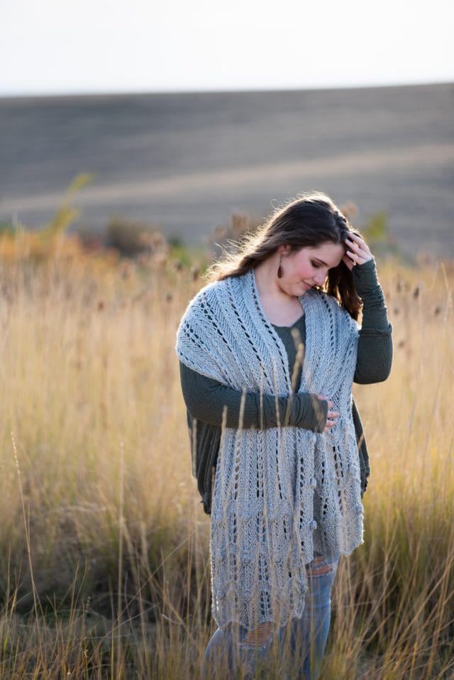 A woman wears an oversized knitted gray shawl/scarf around her neck. Rainsong wrap by Kalurah Hudson, a new indie knitting pattern available at knitpicks.com.
