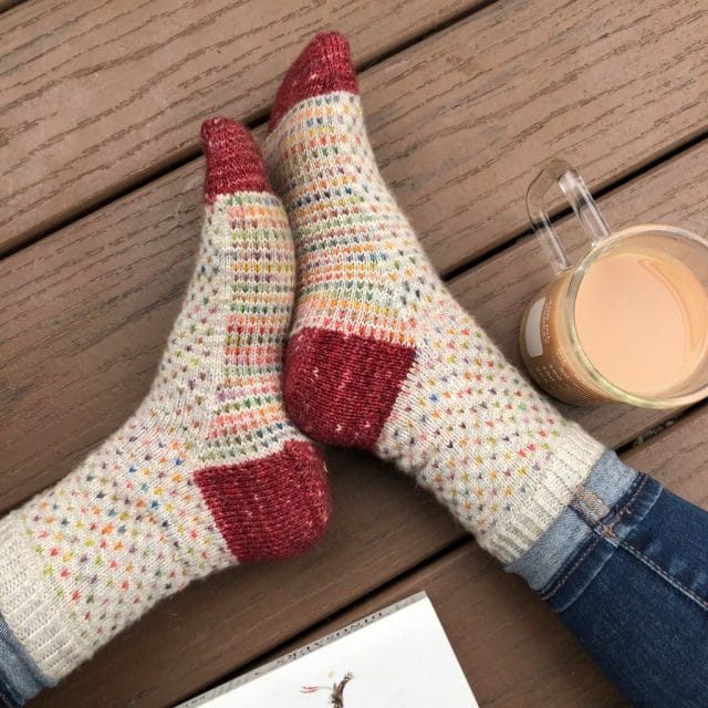Two feet wearing adorable knitted socks featuring an off-white background with multicolored dots, and contrasting red toes and heels. Flea Circus Socks by Belinda Barbagallo, a new indie knitting pattern available at knitpicks.com.