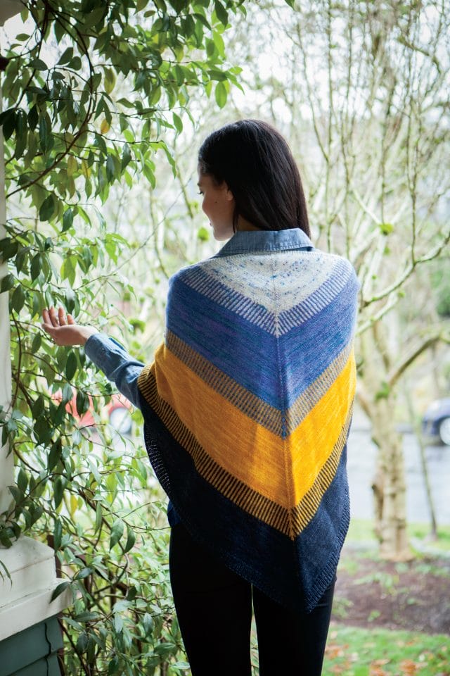 Shown from the back, A model wears a blue + yellow knitted shawl, the Koloreak shawl from Knit Picks.