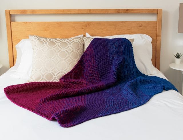 Knit Picks 12 Weeks of Gifting Patterns, featuring the Continuation Blanket. Image shows: a knitted blanket draped on a bed. The blanket gradates from magenta, to purple, to blue, along the diagonal.