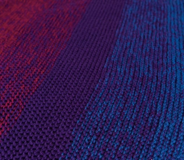 Knit Picks 12 Weeks of Gifting Patterns, featuring the Continuation Blanket. Image shows: a closeup of the knitted texture of the blanket. The blanket gradates from magenta, to purple, to blue, along the diagonal.