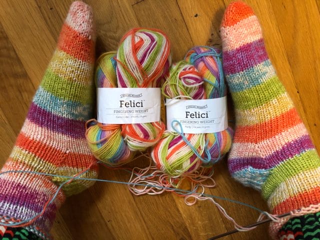 Two feet wearing Rainbow-y knitted socks and several balls of Felici self-striping yarn.