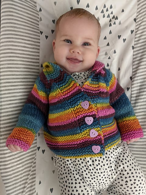 Harvest Sweater by Ravelry user Vyndree. A baby wears a colorful striped knitted cardigan with heart buttons.