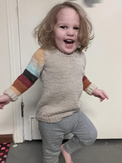 Little worsted Sock Arms by Ravelry user Vyndree. A toddler wears a tan knitted sweater with striped arms.