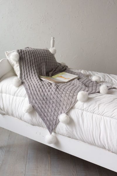 A gray knitted throw with white pom-poms accenting its hem