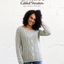 Book cover: text at the top - Chalet, Cabled Sweaters, by Knit Picks - and a photo of a woman modeling a light grey cabled sweater, with columns of cables running up the whole front of the body, including a wider, complex cable in the center, and cables running down the sleeves at the sides only. The woman is smiling and holding her left hand up to her curly hair, standing in front of a white background, and wearing blue jeans.