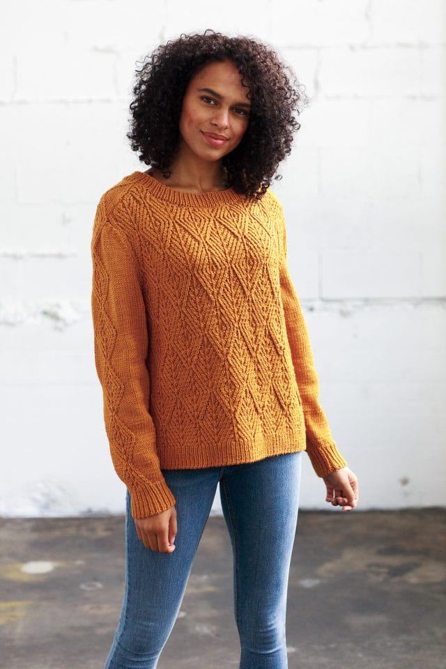 Model wearing a golden yellow pullover, the body completely covered in a repeating diamonds twisted stitch pattern, and a single column of twisted diamonds running up the sleeve and over the shoulder.