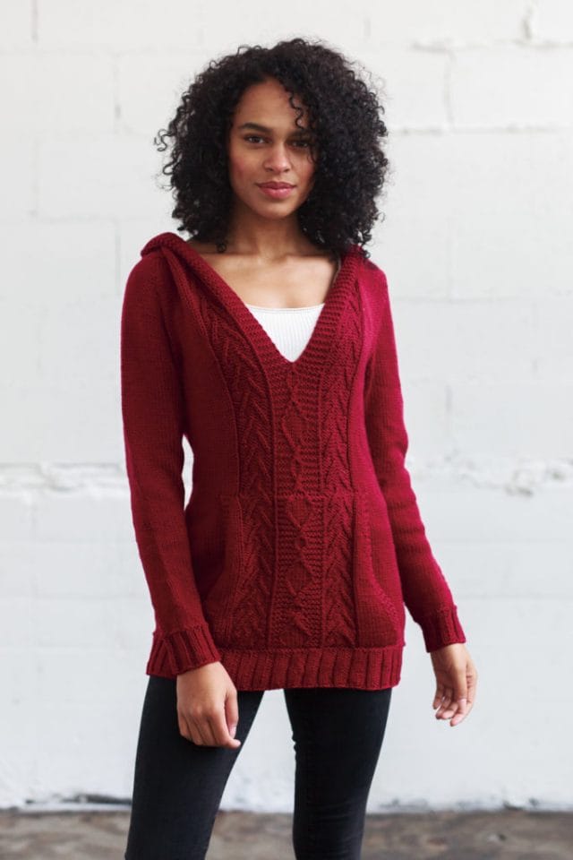 Model wearing a knitted red pullover with deep V-neck and hood in back, and kangaroo style pocket in front. Some small cables run up the center.