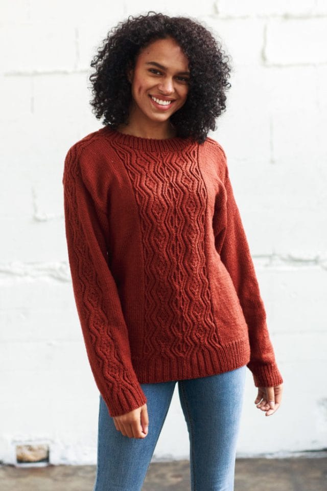 Model wearing a copper-y, orange-ish brown pullover with a wavy cable pattern panel running down from the neckband to the ribbed bottom edge, as well as from the neckband down over the shoulders and down the sleeves to the ribbed bottom.
