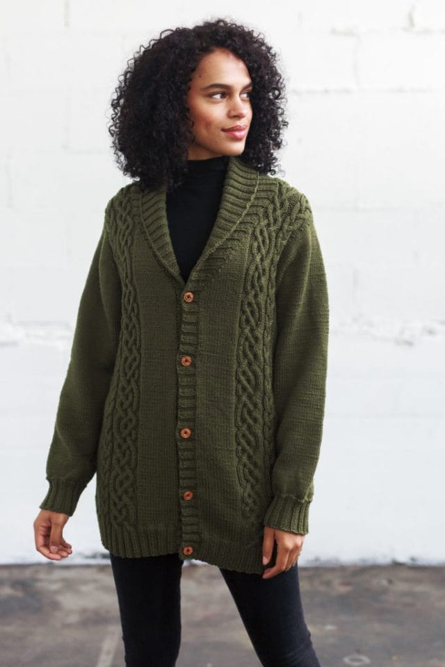 Model wearing mid-thigh length dark green cardigan, buttoned up to a shawl collar, with cables running up the two sides
