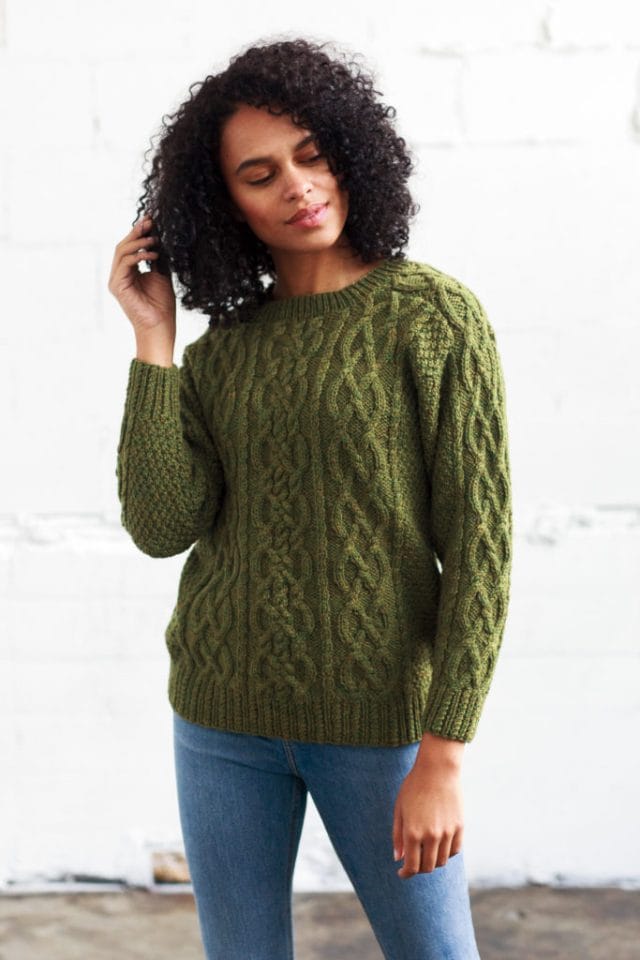 Model wearing a heathered green pullover, covered in cable designs and textured stitches.