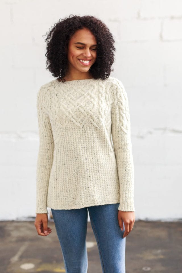 Model wearing white pullover with tweed speckles, cables covered the top of the body and sleeves/shoulders, and a textured rib type pattern covering the middle down to the bottom, of both the body and sleeves.