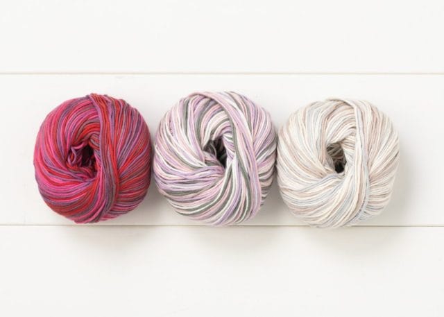 A trio of multi hued cotton yarns on a white wooden background.