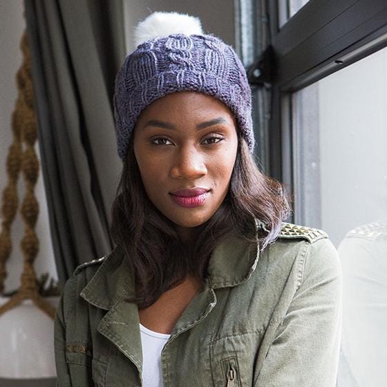 A model wears a purple knitted hat with cables and a white faux fur pom-pom