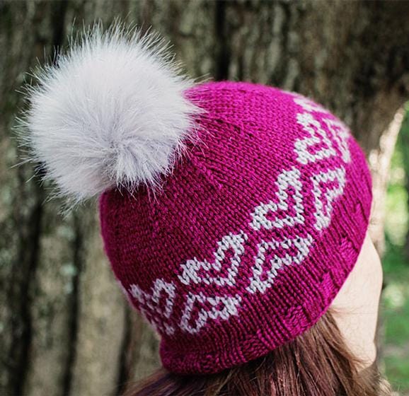 A fuchsia knitted hat with heart motifs and a gray fur pom-pom
