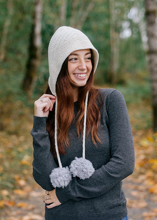 a hood-style knitted hat in white with long strings that end in fur pom-poms
