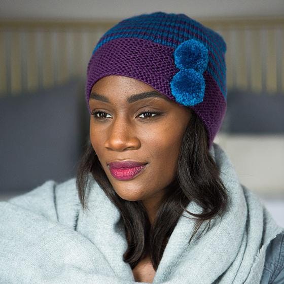 A cloche-style knitted hat with a purple brim accented with two small blue pom-poms