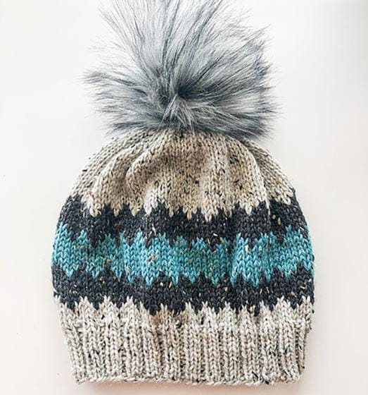 A tweed knitted hat in gray and blue jagged stripes with a large gray fur pom-pom