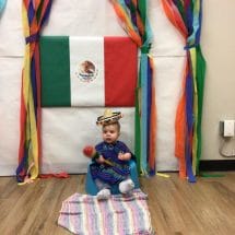 A baby sitting on a blanket on the floor in front of the Mexican flag